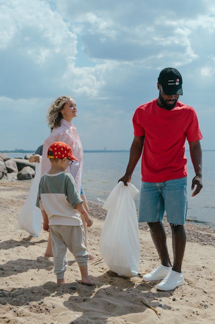 A man wearing a black cap, red T-shirt, ripped denim shorts, and white sneakers is standing on a sandy beach, holding a large white rubbish bag in his right hand, with a stack of rocks and the water visible in the background. Behind him stands a woman with curly blonde hair, wearing a light pink top and dark pants, and a young boy dressed in grey shorts, a T-shirt with green sleeves, and a red cap with yellow patterns, also holding a white rubbish bag. The scene is outdoors near a body of water with partly cloudy skies overhead, suggesting a community or family effort to collect litter from the beach environment, aligning with principles of independent waste collection and on-site rubbish removal, similar to services offered by companies such as Rubbish Clearance Barnet.