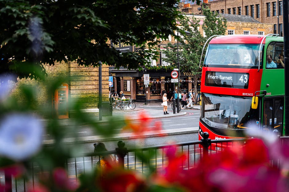 A double-decker bus displaying route number 43 and the destination 'Frieden Barnet' is partially visible on the right side of the image, with its front and side windows showing the driver and passengers inside. In the foreground, out-of-focus colorful flowers and green foliage frame the lower part of the scene. Behind the bus, a busy urban intersection features pedestrians crossing the road, some standing near bicycles parked beside a busy corner. The background reveals a brick building with multiple windows, along with various street signs, including a red and white bus stop sign. Trees with lush green leaves line the street, providing partial shade and adding a natural element to the cityscape. The environment appears lively, capturing a typical day in a vibrant, pedestrian-focused area, consistent with the context of local-level urban rubbish and waste management services like those offered by Rubbish Clearance Barnet, which handle waste collected during city maintenance or private clearance projects.