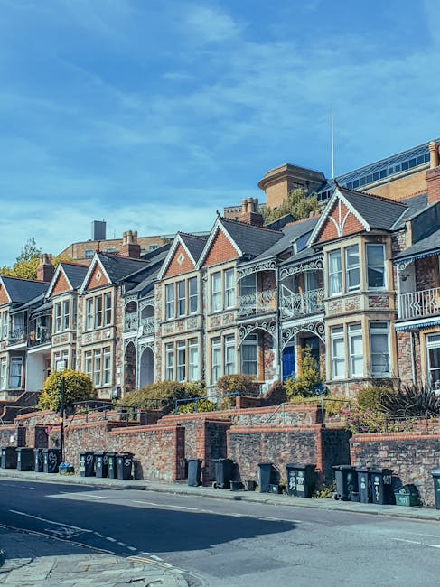 A row of Victorian-style terraced houses with decorative white trim and bay windows, situated along a residential street. The buildings feature brick facades with some painted elements, gabled roofs, and small front gardens enclosed by low brick walls. In the foreground, several black wheelie bins are lined up along the pavement, adjacent to the brick wall boundary of each property. The street appears clean and well-maintained, and the background shows a mix of blue sky with some scattered clouds and modern commercial or office buildings behind the row of residences. The scene suggests a typical urban neighbourhood where private waste collection may take place, aligning with independent rubbish removal services such as those provided by Rubbish Clearance Barnet, supporting local household waste management needs without reliance on local council collection.