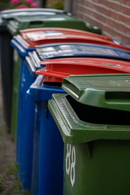 A row of large, rectangular wheelie bins positioned outdoors along a paved pathway adjacent to a brick wall. The bins are made of durable plastic with textured surfaces on the lids and are arranged in an orderly line. The front bin is green with a white recycling symbol on the side, while the other bins behind are blue, red, and dark green, each with matching or contrasting lids. The blue bins appear designated for general waste, and the red bin is likely for other specific waste types. The scene is captured under natural light, with the background slightly out of focus, emphasizing the bins' vivid colors and uniform arrangement. The environment suggests a typical residential or commercial waste disposal setup, possibly associated with independent waste collection services like those provided by Rubbish Clearance Barnet, which specialise in private on-site clearance and rubbish removal outside of local authority collections.