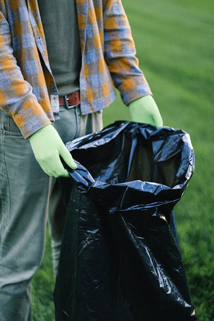 The image shows a person standing outdoors on a grassy area, holding open a black garbage bag with both hands. The person is wearing a plaid shirt with shades of yellow, gray, and blue, along with green rubber gloves, and gray trousers with a brown belt. The black trash bag appears to be empty or ready for disposal, with its top edges folded outward. The background is a lush, green outdoor environment, suggesting a garden or park setting. The scene emphasizes waste collection or rubbish removal, aligning with services such as those provided by Rubbish Clearance Barnet, which specializes in private waste handling and on-site clearance. Natural lighting highlights the textures of the gloves and the crinkled plastic of the bag, while the overall composition maintains a professional, neutral tone focused on environmental and waste management activities.